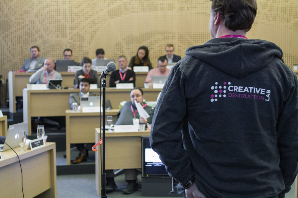 A program leader stands with his back to the camera while beyond him sits a small auditorium of students